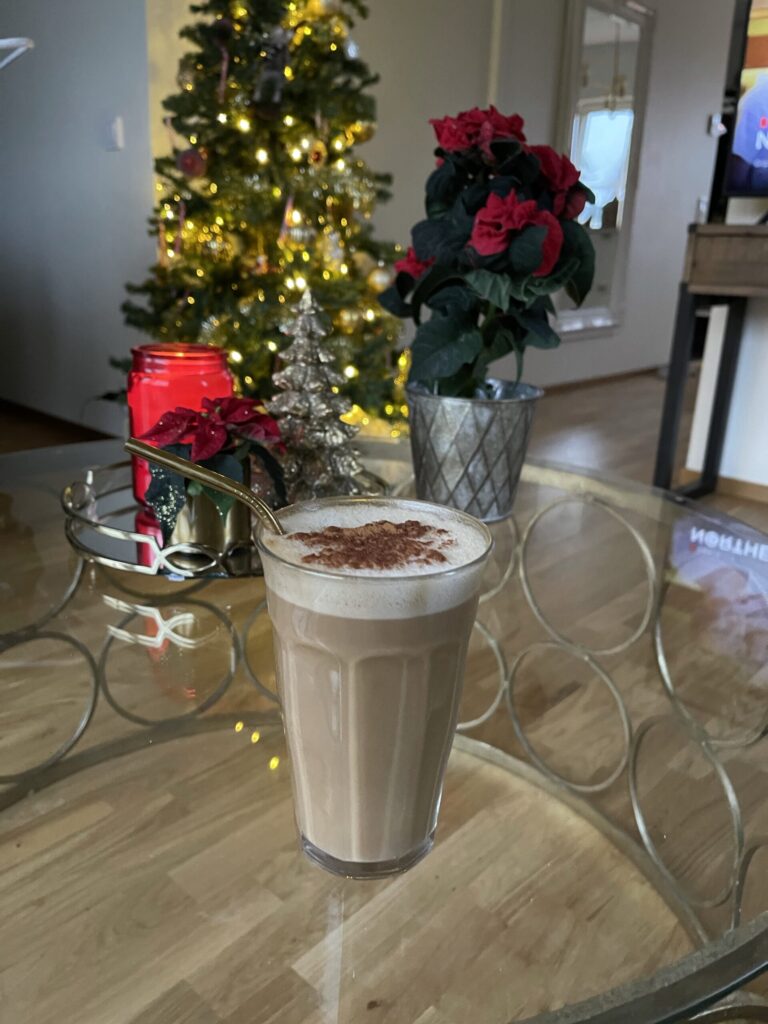 Ett glas står på ett glasbord. I glaset är det kaffe med mjölkskum som är toppad med kanel. I bakgrunden ser man en julgran och en julstjärna. 