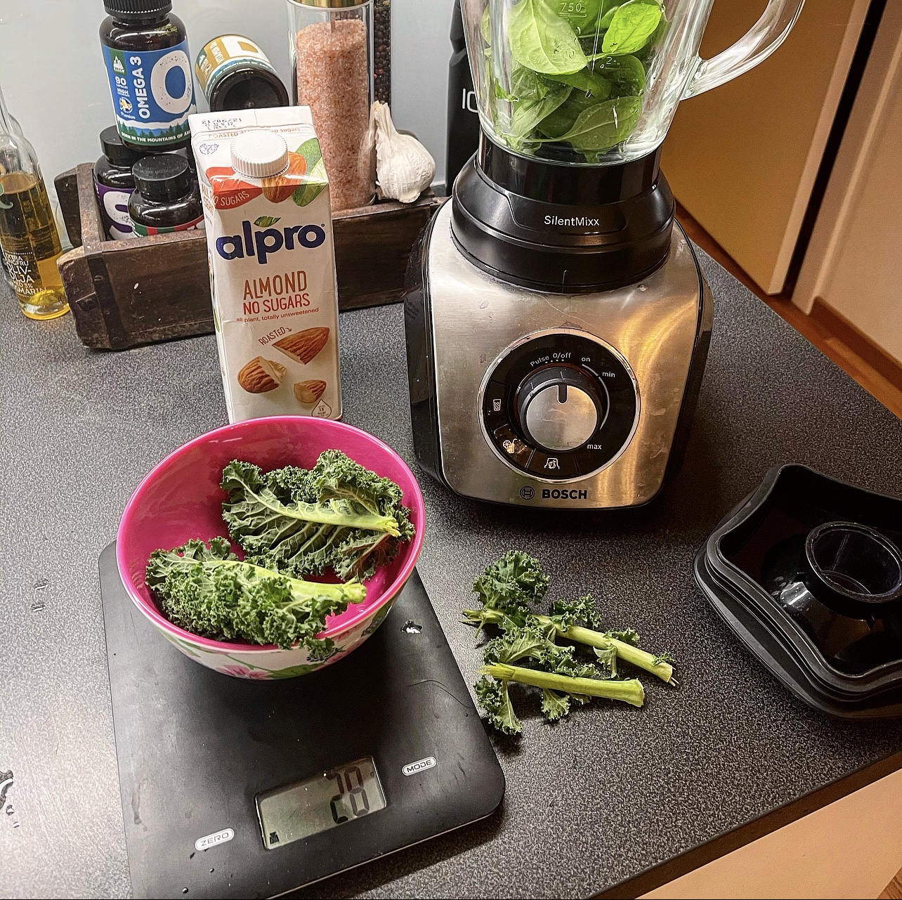 En mixer stör på köksbänken. Förbereder den gröna jäkeln. På bänken står även en matvåg som väger en skål med grönkål. En mandelmöjl från Alpro står även på köksbänken