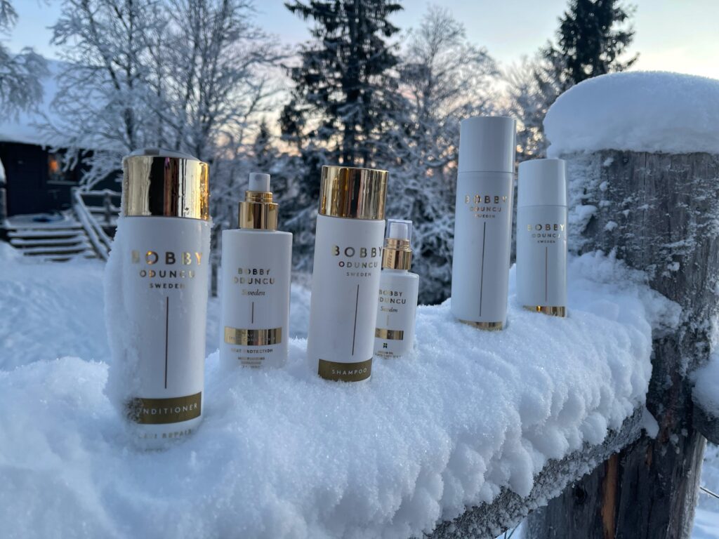 Bobby Oduncu Swedens hårprodukter står på ett räcke täckt i Snö. Grymma produkter för håret. Bakom syns en skog som också är täckt i snö.